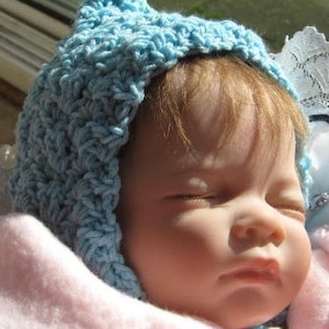 May include: A baby doll wearing a light blue crocheted hat. The doll has closed eyes and a soft, realistic appearance.