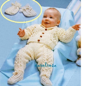 May include: A crocheted baby outfit in white with a cable knit pattern. The outfit includes a cardigan with buttons and matching pants with booties. The outfit is shown on a baby sitting in a chair.
