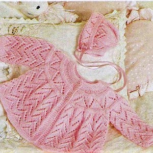 Puede incluir: Conjunto de bebé de punto rosa claro que incluye un cárdigan y un gorro a juego. El cárdigan presenta un diseño calado detallado y mangas largas. El gorro tiene un patrón similar y lazos. El conjunto se muestra sobre una superficie suave y texturizada.