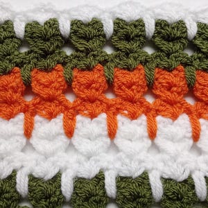 May include: Close-up of a crocheted fabric with a repeating pattern. The design features rows of white, orange, and green yarn, creating a textured and colorful textile. The pattern is intricate and detailed, showcasing the craftsmanship.