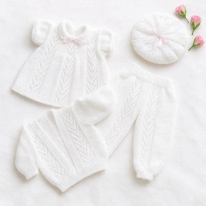 Könnte beinhalten: Ein weißes Strick-Baby-Kleidungsset, bestehend aus einem Kleid mit rosa Schleife, einer Strickjacke, einer Hose und einer Baskenmütze mit rosa Schleife. Das Set wird mit drei rosa Rosen präsentiert.