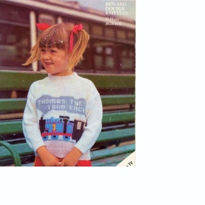 May include: A white knitted sweater with a blue and red Thomas the Tank Engine design. The sweater has the text "Thomas the Tank Engine" on the front. The sweater is being worn by a young child sitting on a bench.