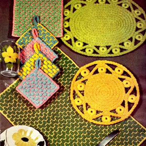 Op de afbeelding: Een verzameling kleurrijke, handgemaakte gehaakte keukenartikelen. Inclusief groene en roze pannenlappen, gele en blauwe pannenlappen, een groen-gele placemat en geel-groene ronde onderzetters. Een glas met een gele bloem is ook zichtbaar.