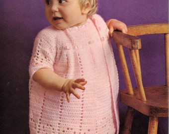 May include: A pink crocheted baby dress with a scalloped hem and a button closure. The dress is worn by a baby girl standing next to a wooden chair.