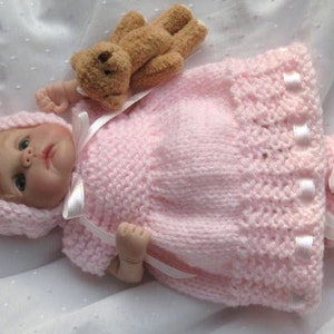 Peut inclure: Une tenue de poupée en tricot rose avec un bonnet et des chaussons assortis. La tenue est ornée de rubans en satin blanc. La poupée tient un ours en peluche brun.