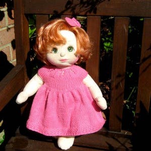 May include: A handmade doll with red hair, wearing a pink knitted dress and a pink flower on her head. The doll is sitting on a wooden chair.