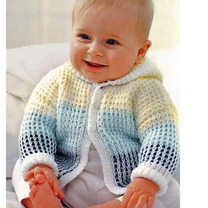 May include: A baby wearing a striped knitted cardigan with a hood. The cardigan is white, yellow, blue, and green. The baby is smiling.