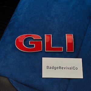 以下が含まれることがあります： 青いテクスチャの表面に、赤とシルバーの「GLI」エンブレム。文字は光沢があり、シルバーの縁取りがあります。「BadgeRevivalCo」と書かれた白い長方形がエンブレムの下にあります。