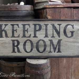 May include: A rectangular wooden sign with the words "KEEPING ROOM" in black, framed by a black border. The sign has a distressed, weathered appearance, suggesting a vintage or rustic style. The background includes wooden barrels and a wooden cabinet.