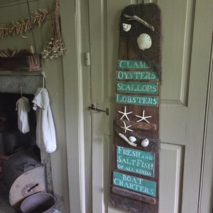 May include: A tall, weathered wooden board hanging on a door, decorated with fishing nets, seashells, and starfish. It features stacked green rectangular signs with white text reading CLAMS, OYSTERS, SCALLOPS, LOBSTERS, FRESH AND SALT FISH OF ALL KINDS, and BOAT CHARTERS.