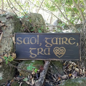 May include: A black wooden sign with gold lettering that reads "saol, gare, gra" with a Celtic knot heart design. The sign is sitting on a pile of rocks in a forest setting.