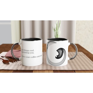 Könnte beinhalten: Weiße Keramik-Kaffeetasse mit schwarzem Innenraum und silberfarbenem Griff. Eine Seite zeigt den Text "All things end. Including you. Have your coffee anyway." Die andere Seite hat ein abstraktes Design in Schwarz und Grau.