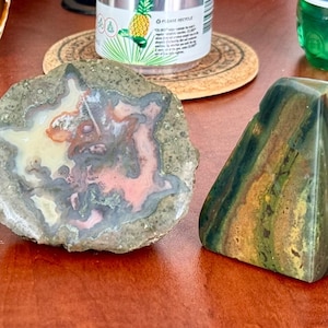 May include: Five polished agate stones in various shapes and colors, including brown, green, red, and white. The stones are arranged on a wooden surface.