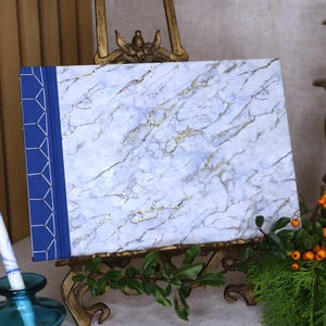 Puede incluir: Un cuaderno con cubierta efecto mármol blanco y azul claro con vetas doradas. Tiene un lomo azul con un patrón geométrico. El cuaderno se exhibe sobre un caballete dorado decorativo.