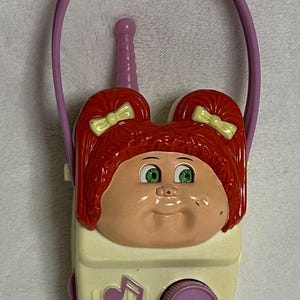 May include: A vintage toy radio featuring a Cabbage Patch Kids design. The radio has a cream-colored body with a purple dial and handle. The face of the radio is a Cabbage Patch Kids doll with red hair, green eyes, and yellow bows. The radio measures approximately 6 inches tall.