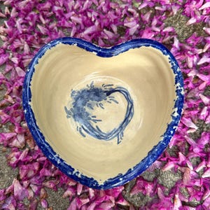 May include: Heart-shaped ceramic bowl with a cream-colored interior and a blue rim. The bowl features a blue floral design in the center. The bowl is surrounded by purple flower petals.