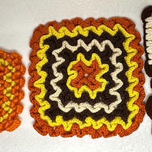 May include: A collection of four handmade crochet squares in warm colors. The squares feature intricate patterns with shades of orange, yellow, brown, and cream. The designs include floral and geometric motifs, perfect for home decor or craft projects.