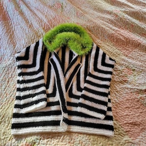 May include: A handmade crochet jacket with a bold black and white striped pattern. The jacket features a vibrant green, fuzzy collar. The garment is displayed on a floral patterned bedspread, showcasing its unique design.