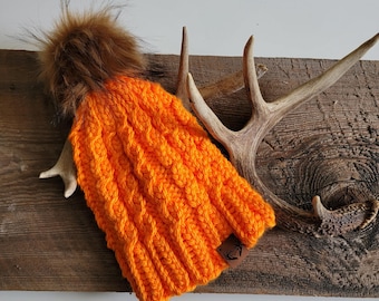 May include: Orange knitted beanie with a brown faux fur pom pom. The beanie has a cable knit pattern and a brown leather patch with a deer logo.