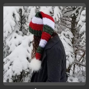 May include: A long, striped knit hat in red, white, and green, with a white pom-pom at the end. The hat is worn by a person in a dark gray coat, set against a snowy forest backdrop. The hat's design is reminiscent of Christmas colors.
