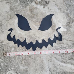 May include: A dark blue Halloween face decal with jagged teeth, large eyes, and a curved mouth. A measuring tape shows the decal is approximately 25 cm wide. The decal is on a clear backing.