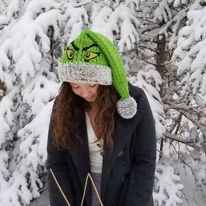 以下が含まれることがあります： 黄色の目と黒い輪郭が特徴の、グリンチの顔のデザインが施された緑色の手編みの帽子。帽子には灰色のフェイクファーのトリムと灰色のポンポンが付いています。背景は雪に覆われた森です。