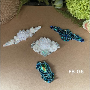 May include: Four decorative embellishments with a variety of designs. Two are white with iridescent crystals and two are teal with iridescent crystals. The teal embellishments have a floral design and the white embellishments have a floral and skull design. The text "FB-G5" is in the lower right corner of the image.