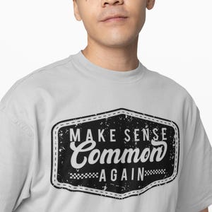 May include: A light gray t-shirt with a black and white graphic that reads "MAKE SENSE Common AGAIN". The graphic is in a distressed style, with a black border and a checkered pattern at the bottom.