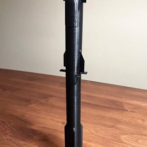 May include: A black 3D-printed model rocket, standing upright on a wooden surface. The rocket features a pointed nose cone, fins, and landing legs. The model is a detailed representation of a spacecraft.