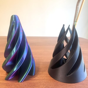 May include: Two abstract, cone-shaped sculptures. One is iridescent with blue, purple, and green hues. The other is solid black. Both have a spiral design with curved, open sections. They sit on a wooden surface.