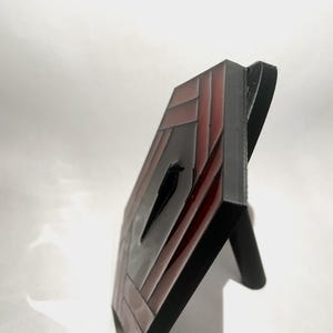 May include: A decorative, geometric art piece with a black bird silhouette. The artwork features a rectangular frame with a red and black striped pattern. The piece is supported by a black stand.