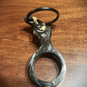 May include: A handcrafted black and gold key ring. The key ring features a textured, organic design with gold leaf accents. The ring itself is a circular shape, and a black metal ring is attached for keys. The key ring is made of a dark material.