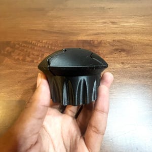 May include: A black bottle stopper with a unique design. The stopper has a rounded top with a series of grooves along the sides. The stopper is held in a hand against a wooden background.