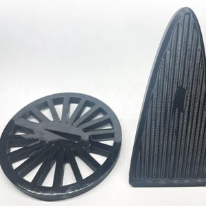 May include: Two black plastic objects on a white background. One is a round, slotted disc. The other is a tall, triangular shape with vertical grooves. Both objects appear to be 3D printed.