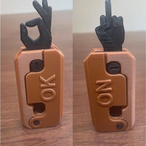 May include: A 3D-printed, two-sided novelty item with a brown and tan case. One side displays an "OK" hand gesture, the other a middle finger. The word "OK" is printed on one side, and "NO" on the other.
