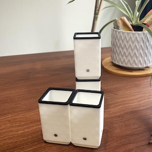 May include: White rectangular planters with black trim. The planters are arranged in a cluster on a wooden surface. One planter is stacked on top of two others. A small, round detail is visible on the front of each planter.