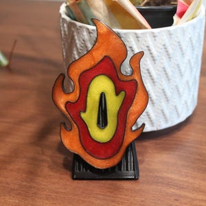 May include: A decorative tsuba sword guard featuring a layered flame design with a shimmering orange outer border, a solid red middle section, and a bright yellow center with a central black vertical opening, mounted on a black stand.