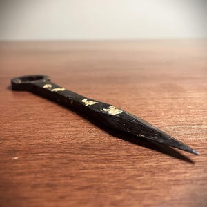 May include: A black metal tool with a pointed end and a circular handle. Gold leaf accents the surface of the tool. The tool is lying on a wooden surface.