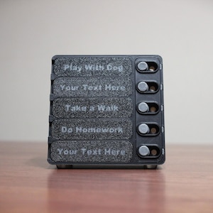 May include: A black, rectangular device with five toggle switches and text labels. The labels read "Play With Dog", "Your Text Here", "Take a Walk", "Do Homework", and "Your Text Here". The device is on a wooden surface.