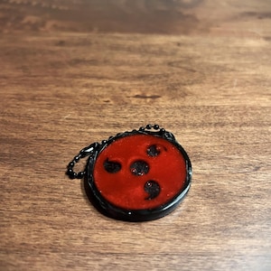 May include: A red and black keychain charm with a circular design. The charm features a red center with three black comma-shaped markings and a black border. A small black chain is attached.