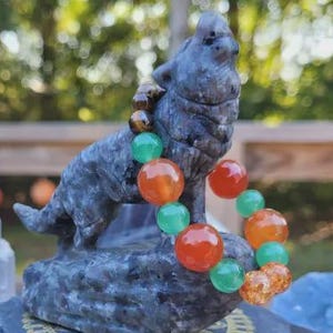 May include: A grey wolf statue wearing a necklace of orange, green, and brown beads. The wolf sits on a dark grey base with a gold pattern. Other crystals and a small carved figure are also present.