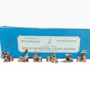 May include: A set of six pewter miniature birthday candle holders featuring characters from The World of Peter Rabbit by Beatrix Potter, displayed in front of a blue box labeled with the product name and Godinger Silver branding.