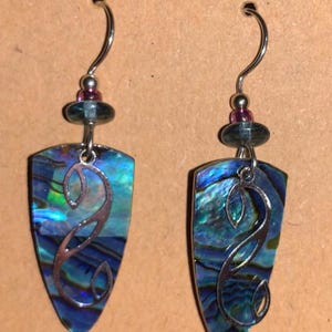 May include: A pair of iridescent abalone shell earrings with a shield shape and silver-tone filigree design. Each earring has a silver hook and is accented with a small purple bead and a dark green bead.