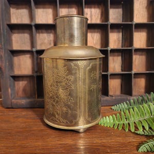 May include: A gold metal tea caddy with a floral design and Chinese characters. The caddy has a lid and a round base.
