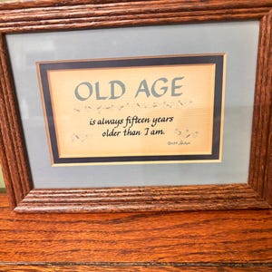 May include: A rectangular wooden frame with a light blue matting surrounding a print. The print features the text OLD AGE is always fifteen years older than I am in dark blue lettering, with small decorative flourishes and a copyright date.
