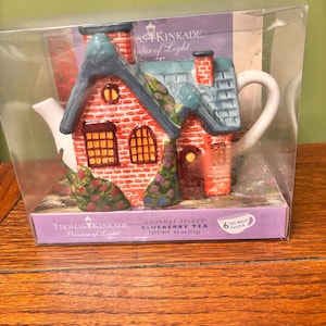 May include: A decorative teapot shaped like a cottage, featuring a red brick exterior, blue roof, and a white handle. The packaging includes the text "Thomas Kinkade Painter of Light" and "Gourmet Select Blueberry Tea, Net Wt. 42 oz (11g)."