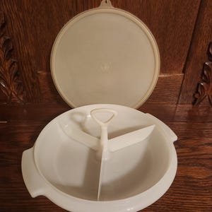 May include: A white plastic serving dish with a lid. The dish is divided into three sections and has a handle in the center. The lid is round and has a small handle. The dish is designed for serving food.