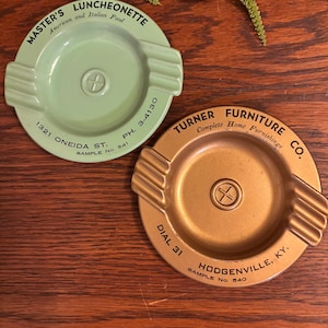 May include: Two vintage ashtrays, one green and one gold, with advertising for a luncheonette and a furniture company. The green ashtray has the text "MASTER'S LUNCHEONETTE American and Italian Food 1321 ONEIDA ST. PH. 3-4130 SAMPLE NO. 541" and the gold ashtray has the text "TURNER FURNITURE CO. Complete Home Furnishings DIAL 31 HODGENVILLE, KY. SAMPLE No. 840".
