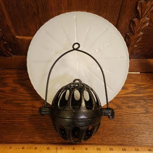 May include: An antique black metal hanging lantern with a white glass shade. The lantern has a spherical cage design and a handle. The shade is a large, round, white glass plate. A wooden ruler is at the bottom.