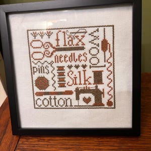 May include: Framed cross-stitch art featuring brown text and illustrations of sewing tools and materials. Words include "flax," "needles," "pins," "wool," "silk," and "cotton." The artwork is in a black frame.